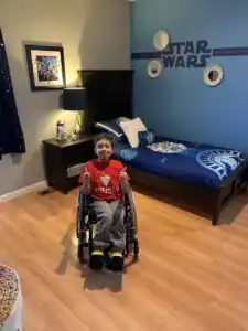 A boy in a wheelchair smiles and gives a thumbs-up in a bedroom decorated with a Star Wars theme, featuring blue walls and a Millennium Falcon bedspread.