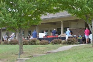 People gather outdoors near a building with tables set up under a covered patio. Some are sitting while others stand and talk. Trees and bushes are in the foreground.
