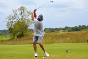 A person wearing a white shirt, gray shorts, and a cap is swinging a golf club on a green golf course with trees and grass in the background.