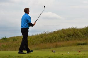 A man in a blue shirt and black pants is standing on a golf course, holding a golf club after taking a shot.