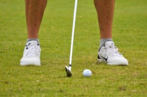 A person stands on grass preparing to hit a golf ball with a golf club; only their legs, feet, club, and ball are visible.
