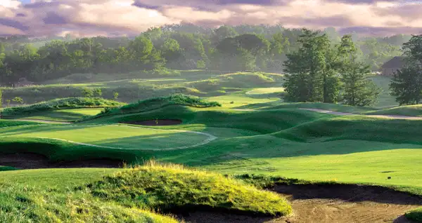 A scenic golf course with rolling green hills, sand bunkers, and tree-lined fairways under a partially cloudy sky.