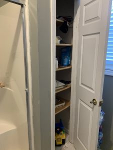 A small bathroom closet with open shelves containing assorted items, next to a shower and a closed white door with a gold handle.