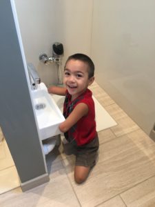 A young boy with two arm differences is kneeling in front of a small sink in a restroom, smiling at the camera.
