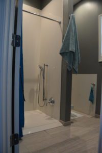Accessible walk-in shower with handheld showerhead, a towel hanging on a wall hook, and tiled floor, viewed from an open doorway.