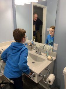 A boy in a blue hoodie looks at himself in the bathroom mirror while an adult stands in the doorway behind him.
