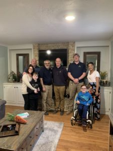 A group of seven people, including a child in a wheelchair, pose and smile for a photo in a living room with a wooden floor and a fireplace in the background.