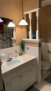 Bathroom with white vanity, blue sink, mirror, overhead lights, partial white divider with columns, and a toilet in the background.
