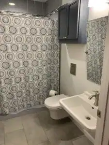 Small bathroom with a patterned shower curtain, wall-mounted toilet, rectangular sink, mirror, and blue wall cabinet above the toilet.