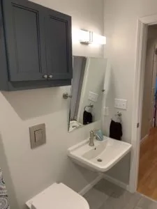 A small bathroom with a wall-mounted sink, a slanted rectangular mirror, a gray cabinet, and a toilet next to a light colored door.