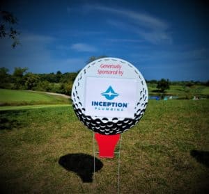 A golf ball-shaped sign on grass reads "Generously Sponsored by Inception Plumbing" with a blue sky and green landscape in the background.