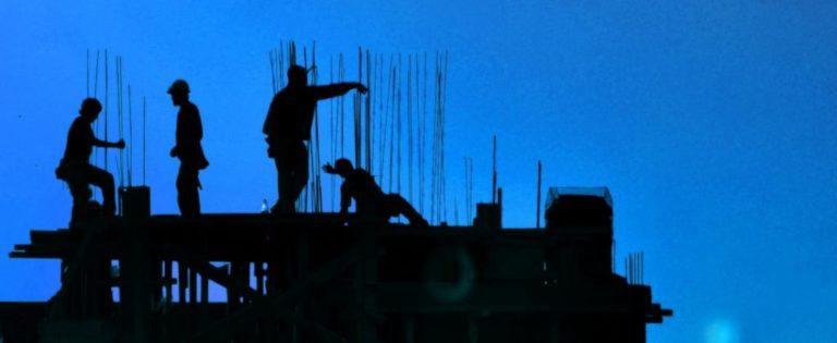 Silhouettes of three construction workers on a building structure with metal rods against a blue sky.