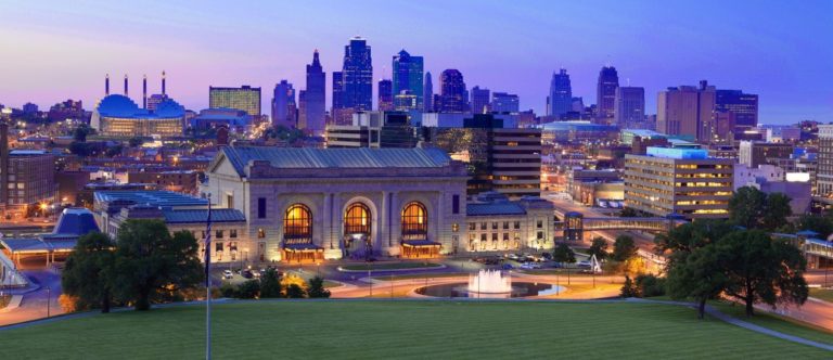 Kansas City skyline at dusk with Union Station in the foreground, illuminated buildings, and a large fountain in front of the station.