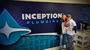 Two people stand in front of an "Inception Plumbing" sign; one holds a bouquet of flowers and a card, celebrating new careers with the company.