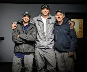 Three men in casual work clothes and hats stand indoors, smiling with arms around each other, in front of a gray wall and a door—celebrating their careers together.