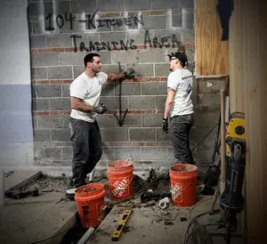 Two construction workers stand in a partially demolished room labeled "104 Kitchen Training Area," with buckets, tools, and debris scattered on the floor—a glimpse into skilled jobs and rewarding careers in construction.