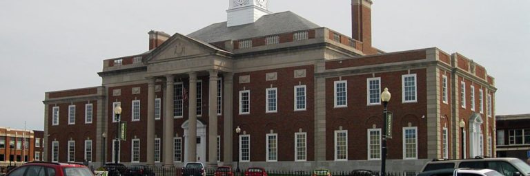 A large brick and stone government building with columns, a clock tower, and parked cars in front.