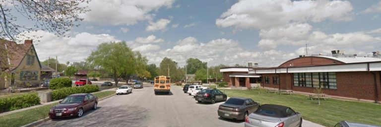 A parking lot with several cars and a yellow school bus sits next to a brick building under a partly cloudy sky. A house and trees are visible on the left.