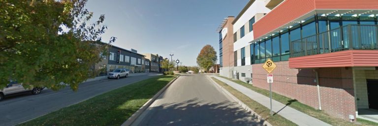 A street runs between modern buildings with a tree on the left and a yellow caution sign on the right under a clear blue sky.