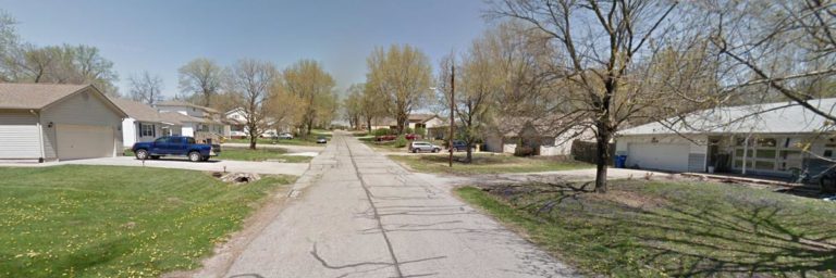 A quiet suburban street lined with houses, trees, parked cars, and a clear sky overhead.