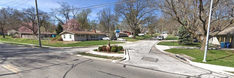 A suburban residential intersection with single-story houses, leafless trees, and a small landscaped traffic island under clear skies.
