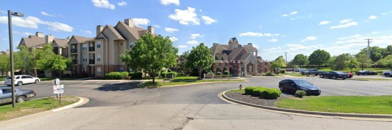A residential apartment complex with multiple buildings, trees, a main entrance, and a parking lot under a blue sky with scattered clouds.