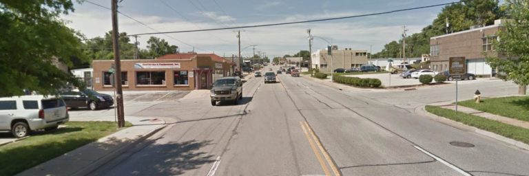 A wide, two-lane street lined with cars, small businesses, and trees on a clear day; traffic is moderate with vehicles in both directions.