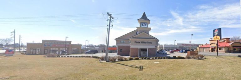 A small clock tower building labeled "Plaza Valley Bank" stands near a street, with businesses and a fast food restaurant visible in the background.