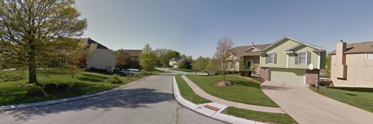 A suburban neighborhood street with single-family homes, green lawns, and trees under a clear blue sky.