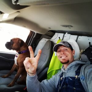 A man in overalls and a cap flashes a peace sign while sitting in a vehicle next to a brown dog in the passenger seat.