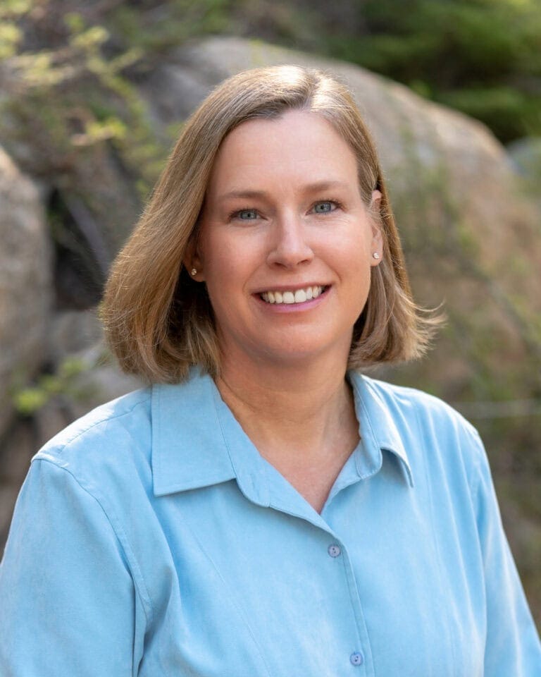 Elegant Denise Bremer smiling, outdoor headshot by water or rocks, natural light, professional image.