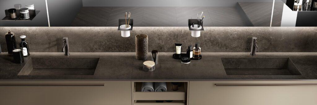 Modern bathroom countertop with two sinks, various skincare and grooming products, and toothbrush holders, against a stone-textured backdrop.