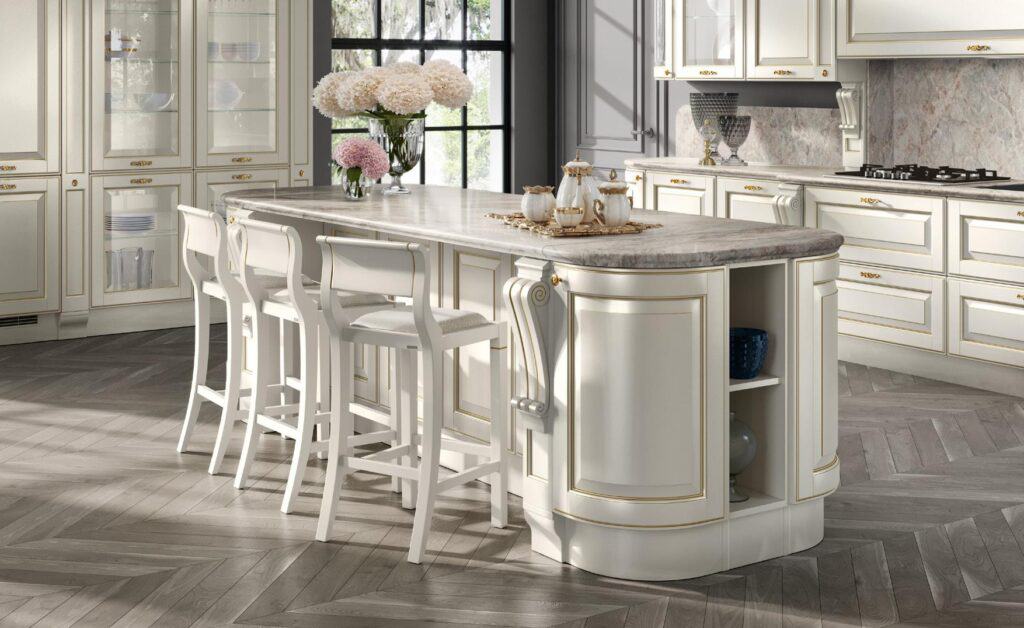 Elegant kitchen with a marble-topped island, white cabinetry, gold accents, and bar stools, featuring floral arrangements and a window in the background.