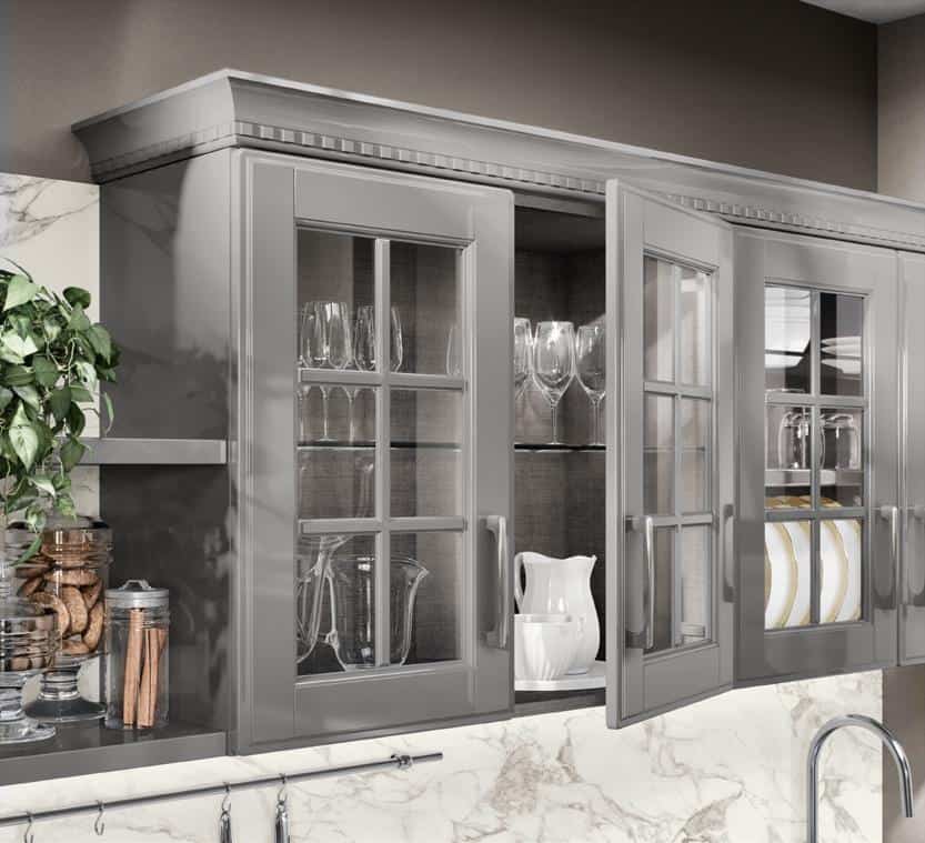 Glass cabinet doors revealing glassware and dishes, with a plant and jars on the left, set against a marble backsplash.