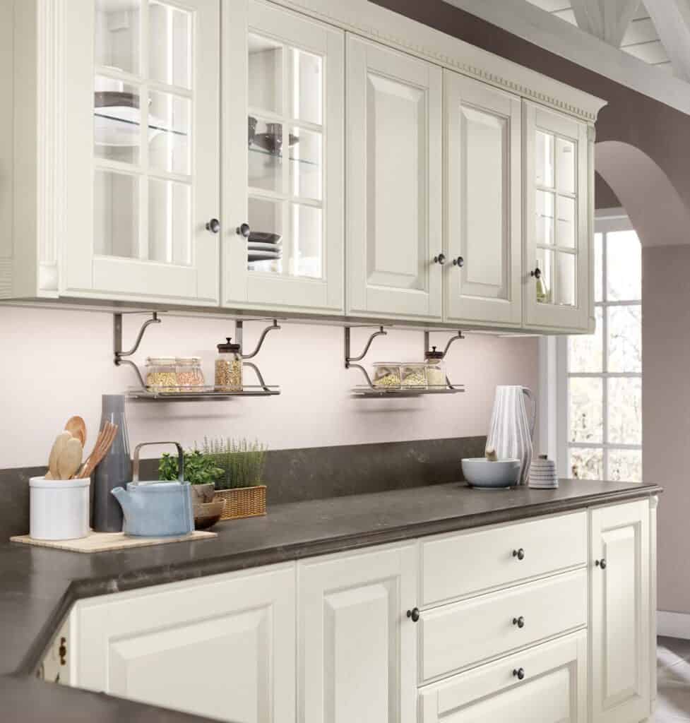 A kitchen with cream-colored cabinets, glass-paneled doors, dark countertops, and a window in the background.