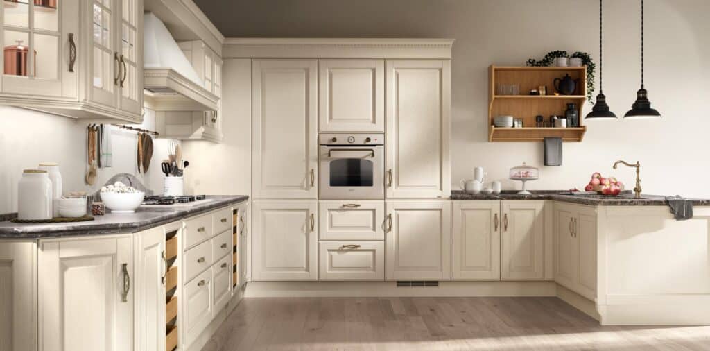 Spacious kitchen with cream cabinetry, dark countertops, built-in oven, and wooden open shelving. Hanging pendant lights illuminate a countertop with a cake stand and fruit.