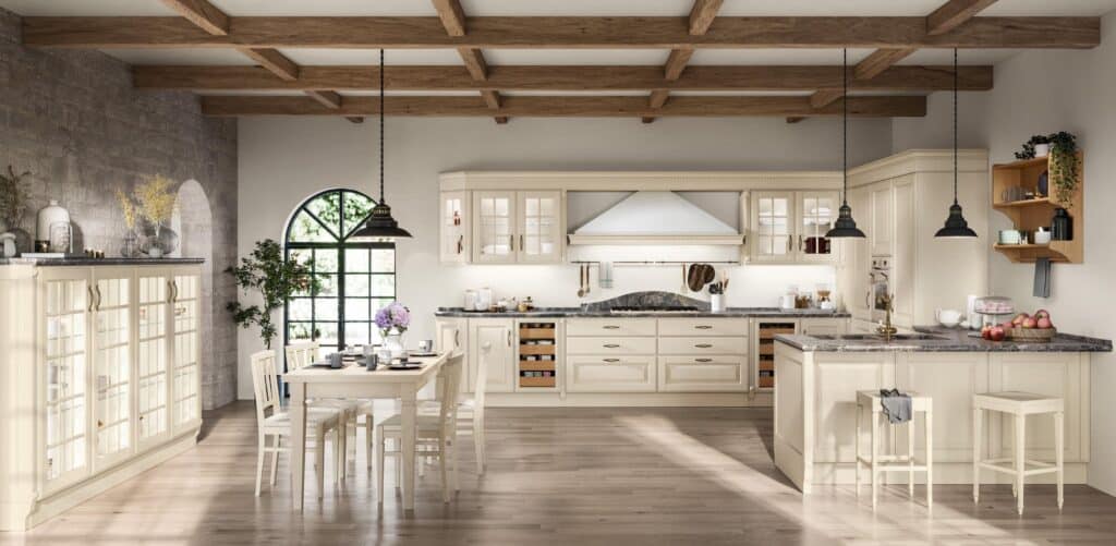 Spacious kitchen with cream-colored cabinets, a large dining table, pendant lights, and a stone countertop under wooden ceiling beams. Large window with greenery outside.