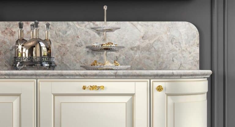 Cream-colored kitchen cabinets with gold handles, a marble backsplash, a tiered dessert stand, and a polished metal kitchen appliance on the counter.