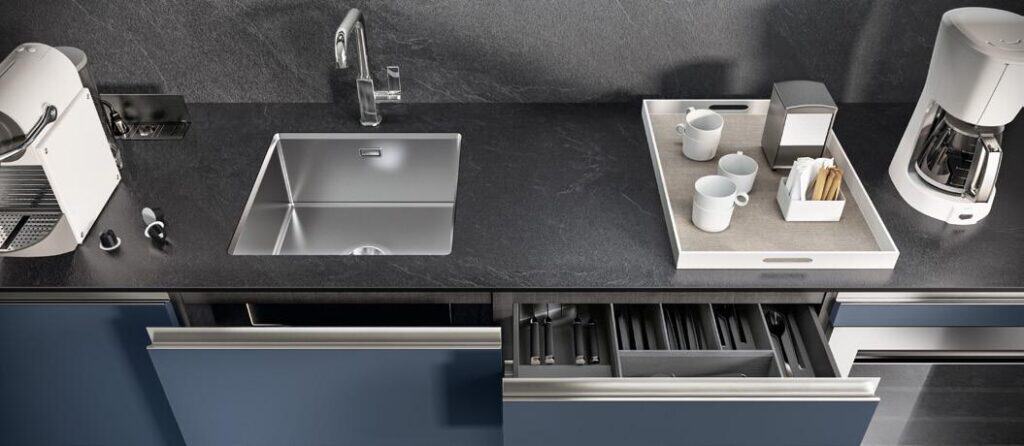 Modern kitchen countertop with a sink, a coffee machine, a tray holding cups and napkins, and an open drawer with utensils.