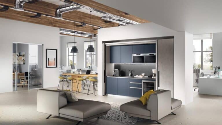 Modern open-concept office with stylish lounge seating, a compact kitchen, large windows, and exposed ductwork.