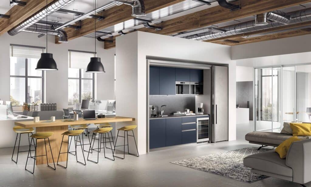 Modern open-plan office space with a kitchen area, large wooden table with bar stools, and a seating area with a sofa.