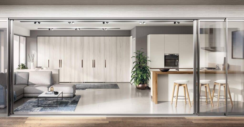 Modern open-plan living room and kitchen with a large L-shaped sofa, a glass coffee table, wood and white kitchen island with bar stools, and sliding glass doors.