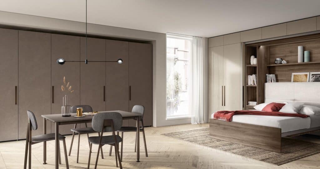 Modern bedroom with a wall of sleek cabinets, a dining table with chairs, and a bed with bookshelves integrated into the headboard. A large window lets in natural light.