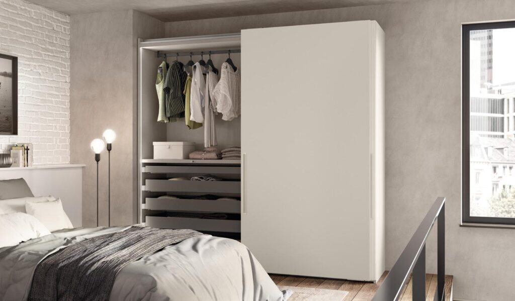 A modern bedroom with a bed and open wardrobe, displaying hanging clothes, shelves, and a window with a city view.