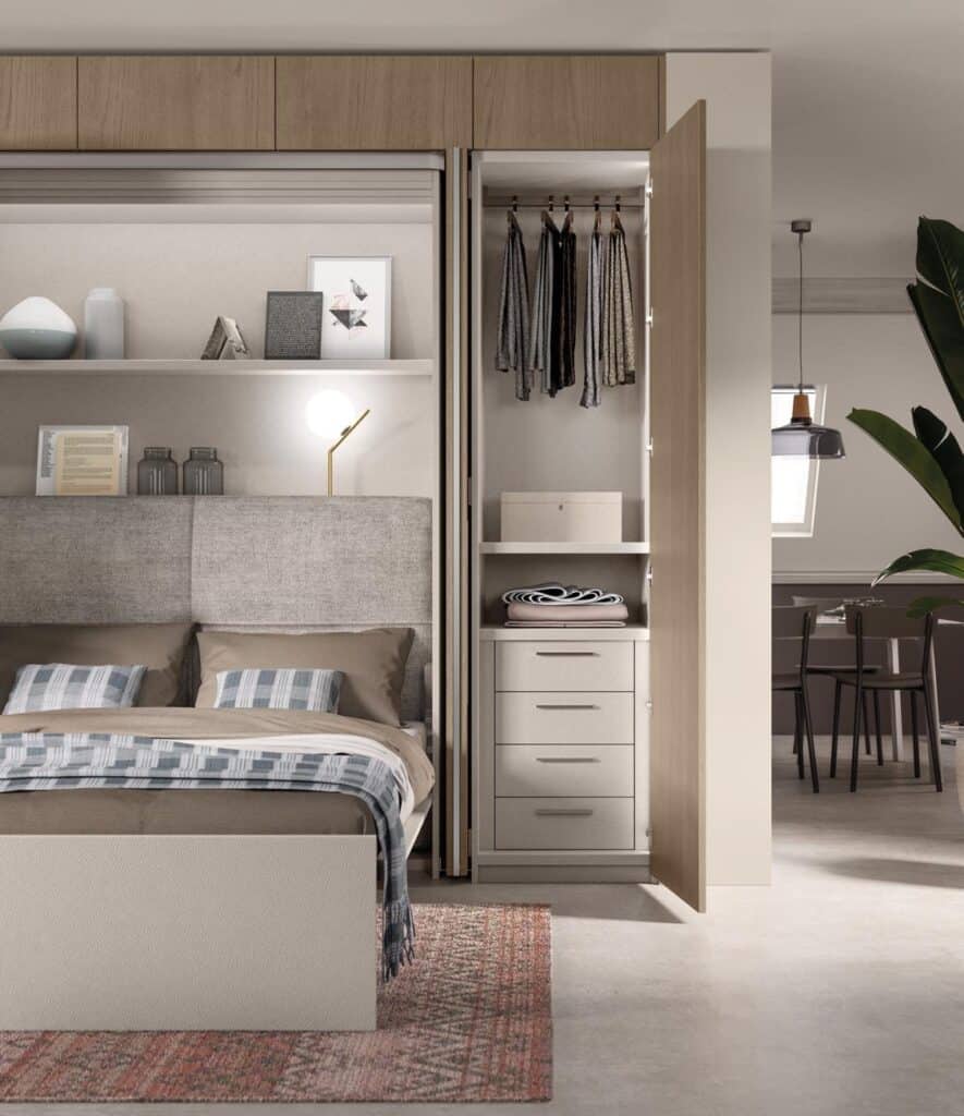 Bedroom interior with a bed featuring beige and plaid bedding, an open wardrobe with hanging clothes, shelves with folded items, and a lamp on a shelf above the bed.