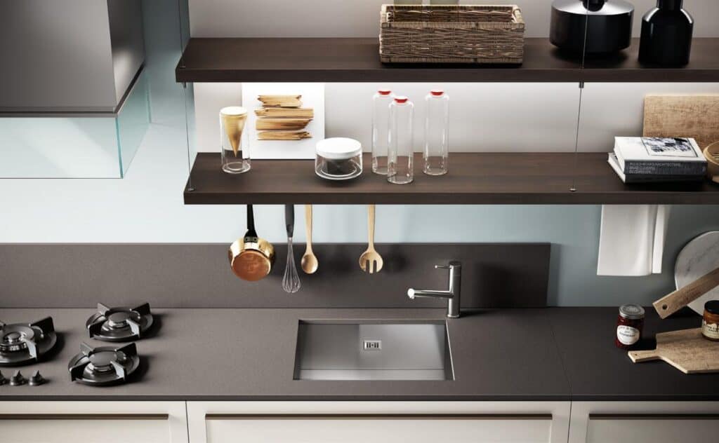 Modern kitchen interior with a built-in cooktop, stainless steel sink, and dark countertop. Wooden shelves hold kitchen utensils, glass bottles, and various decorative items.