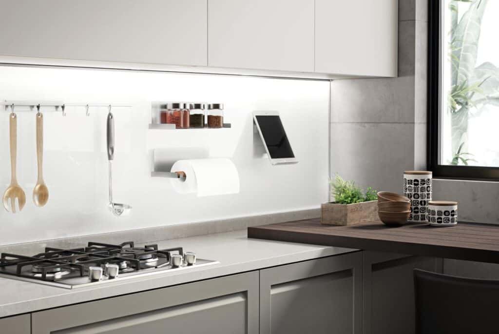 Modern kitchen with a gas stove, wall-mounted spoon and ladle, spices in jars, a paper towel holder, and a tablet on a stand. Countertop accessories include decorative bowls and canisters next to a small indoor plant.