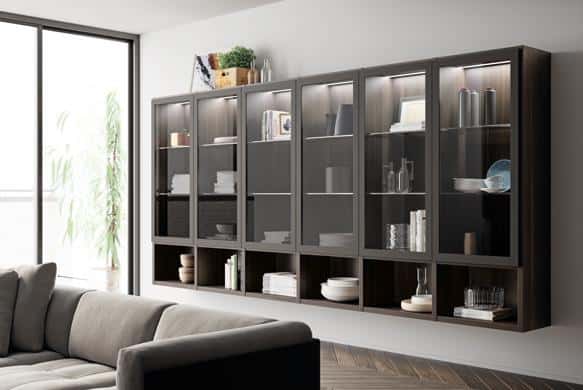 Modern living room with a wall-mounted glass cabinet displaying dishes and glassware, next to a gray sofa and large window.
