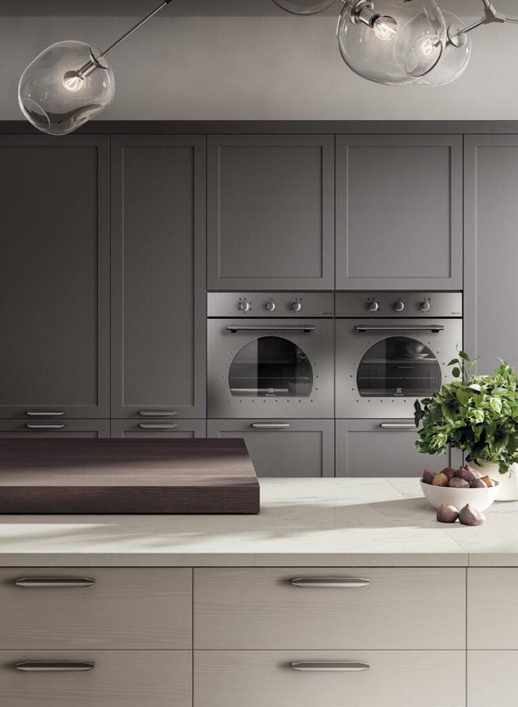 Modern kitchen with gray cabinets, stainless steel double ovens, pendant lighting, and a countertop with a bowl of fruits and leafy greens.