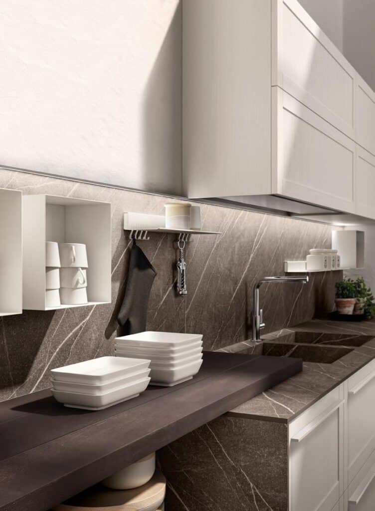 Modern kitchen with dark marble countertops, white cabinetry, stacked white dishes, stainless steel faucet, and small plants.